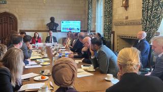 participants of a roundtable breakfast meeting with a tv in the background showing a presentation and the names of speakers 