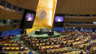 Wide shot shows UN General Assembly Hall, with rows of seated delegates and the President of the General Assembly speaking from the platform